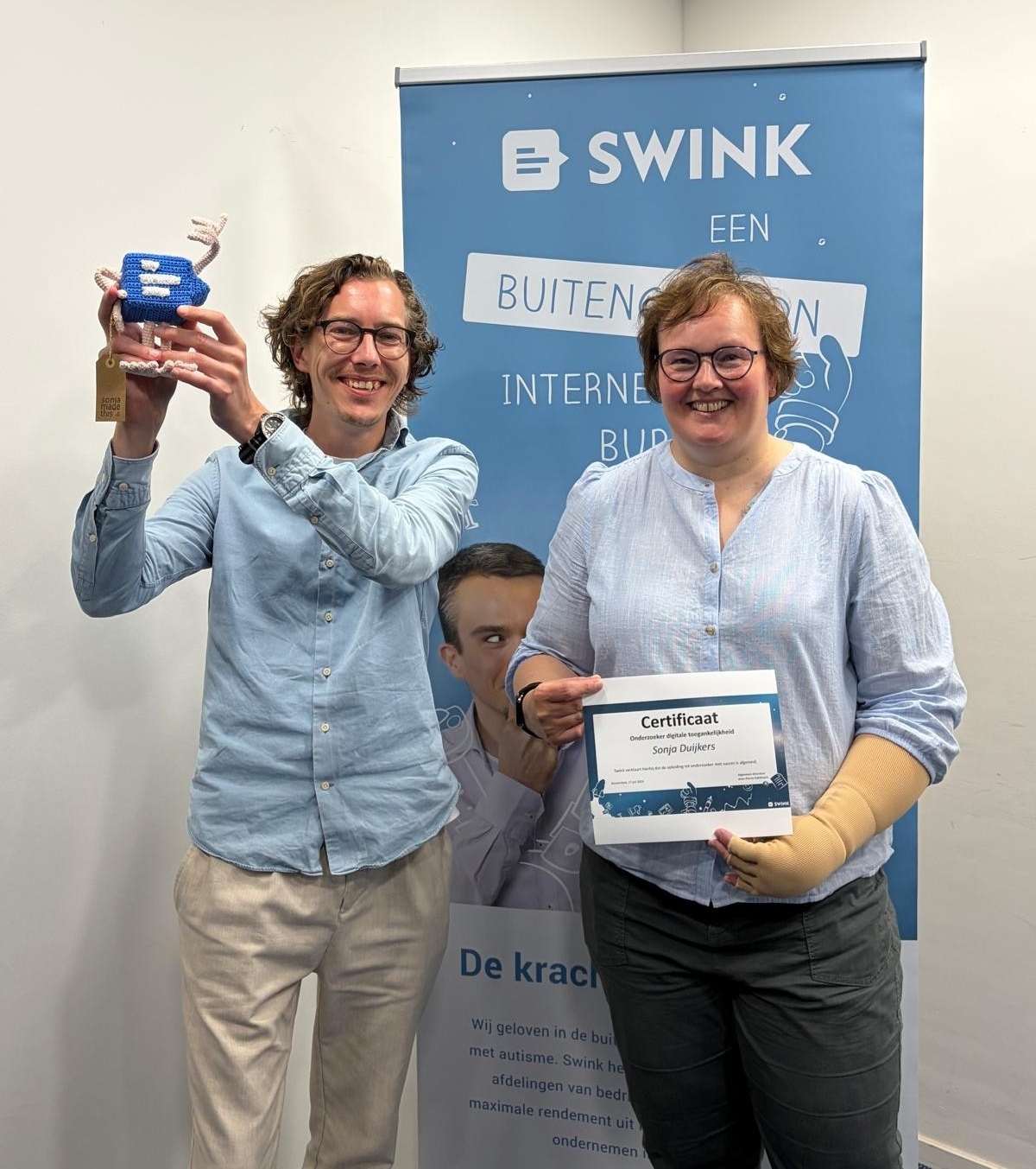 Kenny Heessels en Sonja Duijkers staan voor een Swink banner. Kenny houdt een poppetje van een Swink logo vast.
